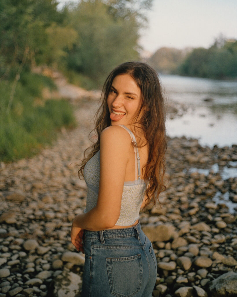 junge frau an fluss schaut über ihre schulter und zeigt zunge analoge farb fotografie greenlight videoproduktion